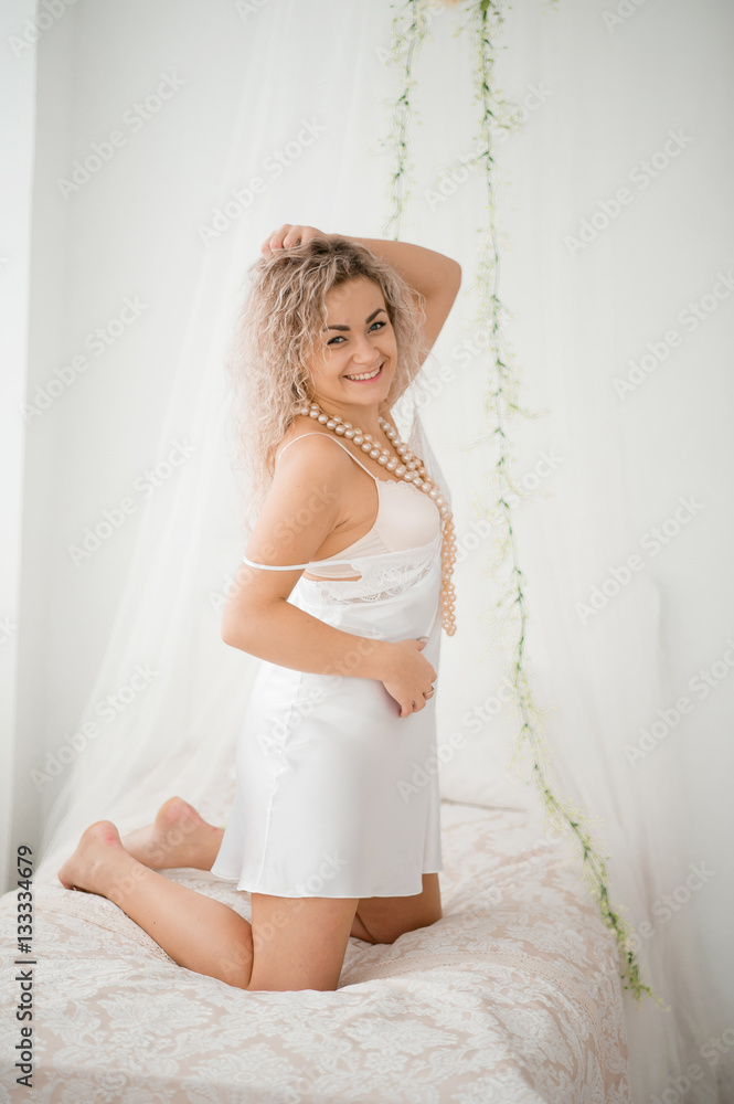 Curly blonde girl in a white silk gown boudoir robe on bed. Curly blonde girl in a white silk gown boudoir robe on bed.