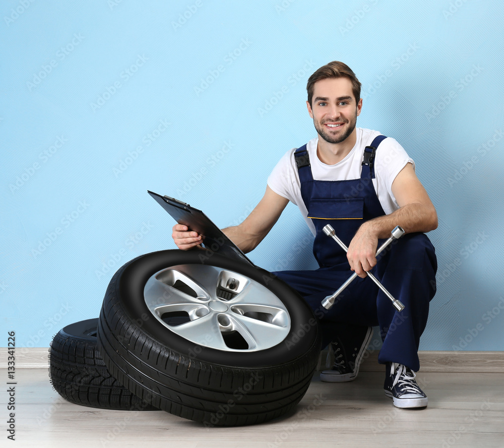 Young mechanic in uniform with spanner and wheels, on blue background Young mechanic in uniform with spanner and wheels, on blue background