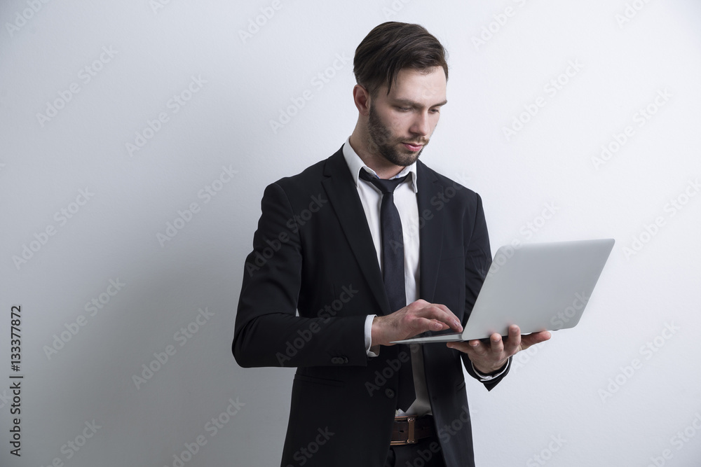 Portrait of a serious bearded young businessman standing near a white wall and working with his laptop. Mock up. Portrait of a serious bearded young businessman standing near a white wall and working with his laptop. Mock up.