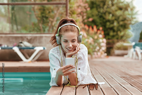 Happy smartphone woman relaxing near swimming pool listening wit