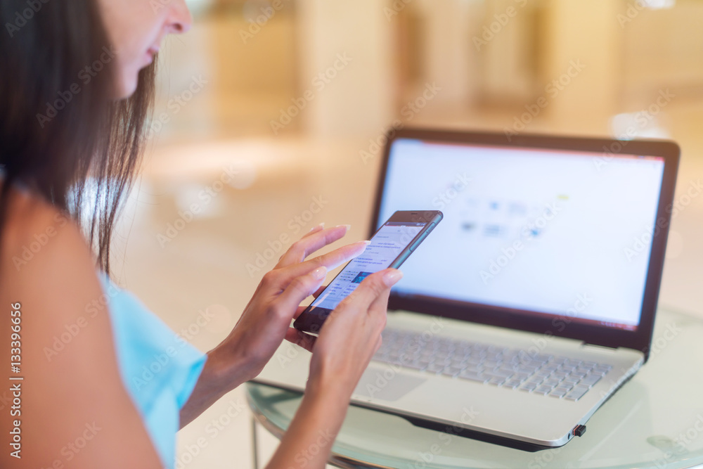 Close-up photo of female hands holding a smartphone with computer laptop in background. Young woman working on cellphone and netbook indoors Close-up photo of female hands holding a smartphone with computer laptop in background. Young woman working on cellphone and netbook indoors