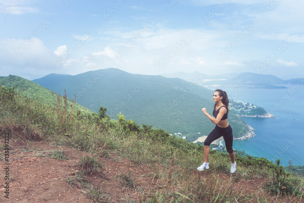 Slender young female athlete doing cardio exercise going up the mountain with sea in background.