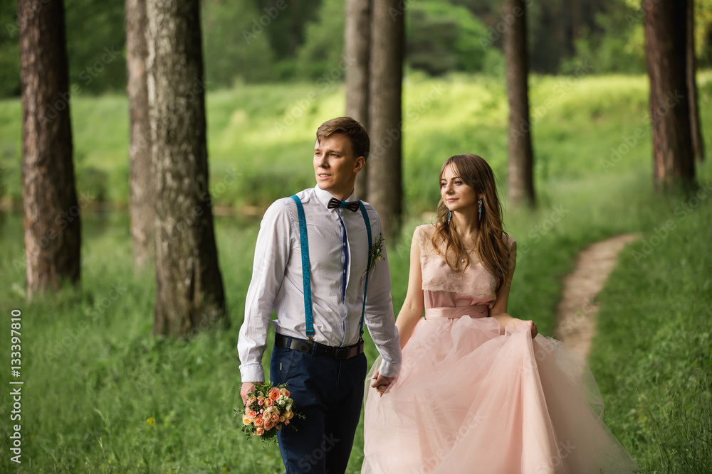 Wedding couple, beautiful bride in peach dress and groom with bow tie and suspenders walking in forest near place of their wedding ceremony