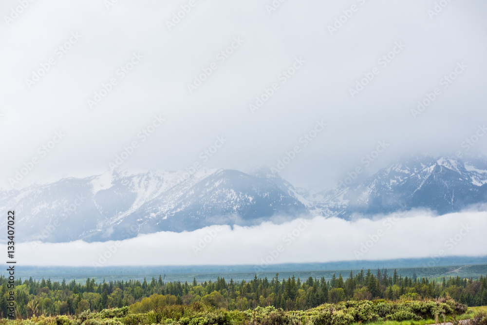 Grand Teton Mountains covered by large storm cloud casting dark shadow on lake with steam geysers