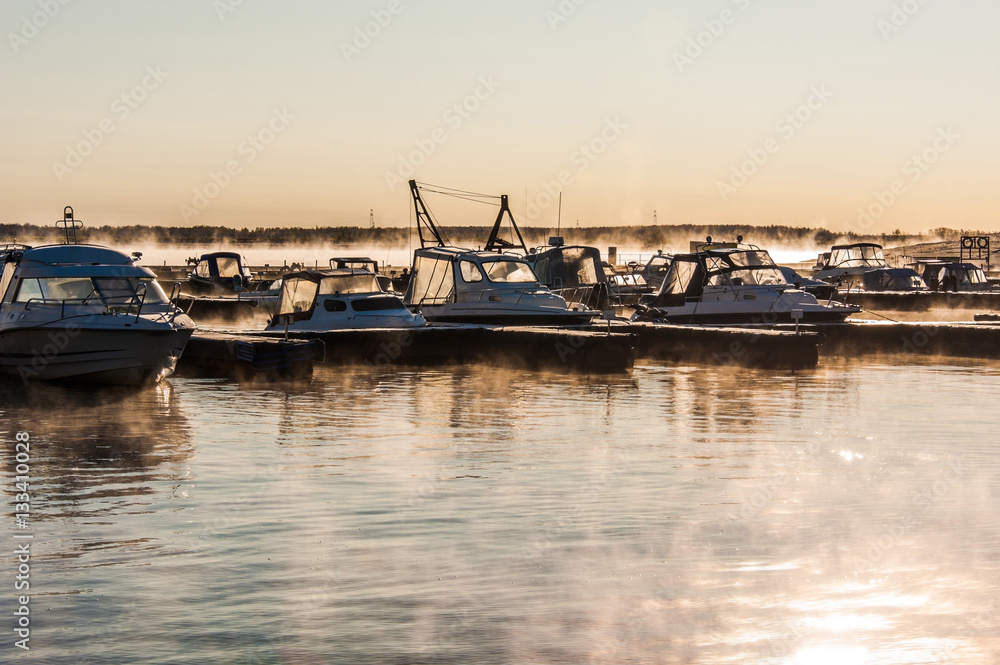 Boats and yachts moored at the port. Glittering sea surface against sunrise. Morning fog. 