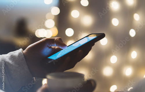 Hipster girl using mobile phone in a home atmosphere, person holding smartphone on background glow bokeh Christmas illimination, female hands texting on relax glitter, mockup templates, blur photo