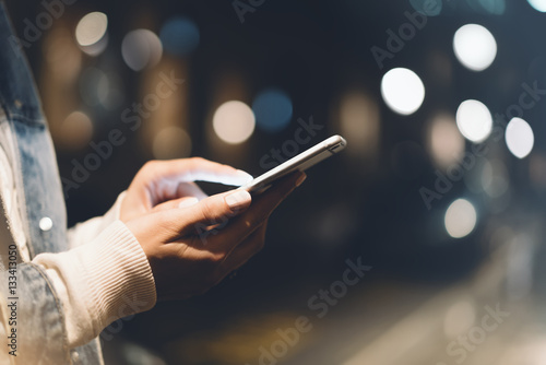 Girl pointing finger on screen smartphone on background illumination bokeh light in night atmospheric city, hipster using in female hands and texting mobile phone, mockup glitter street, content 