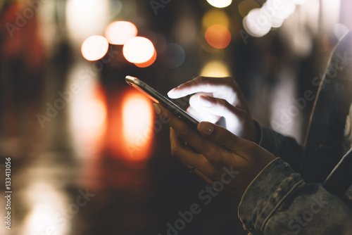 Girl pointing finger on screen smartphone on background illumination bokeh color light in night atmospheric city, hipster using hands and texting mobile phone, mockup glitter taxi street, content 