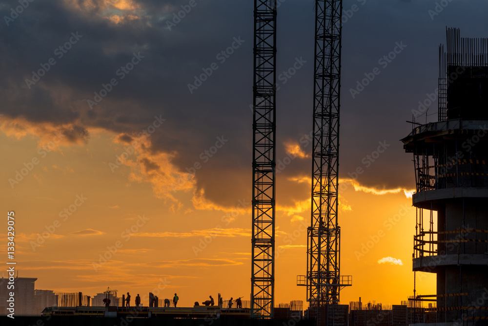 Construction workers silhouettes, cranes and building construction site against sunset sky.