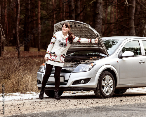 young woman in a white jacket standing near the broken car and asks for help raising thumb up © slavonstok