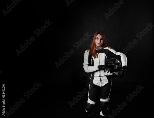 Extreme young girl with red hair in protection body armor of motorcycle sport with helmet in hands. Tired woman after the race on black background with copy space for advertising text.