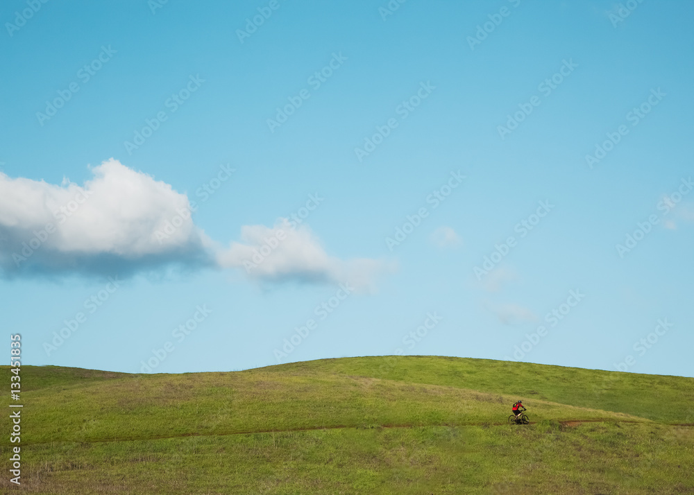Man riding a bicycle on a grassy hill against a blue sky.