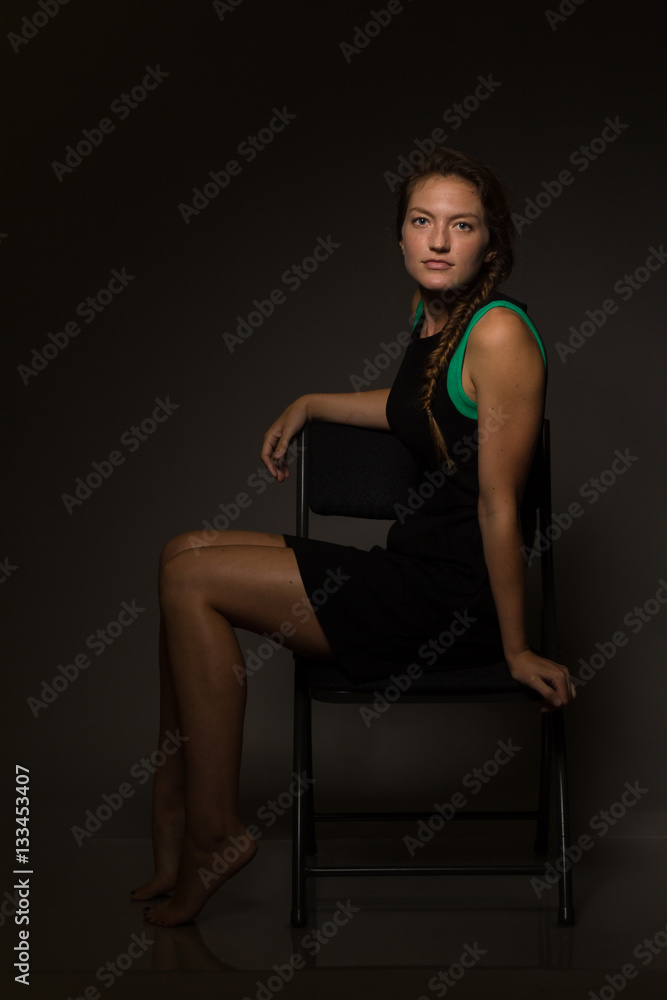 Fototapeta premium Young caucasian woman in a short black dress sits on a chair under moody light