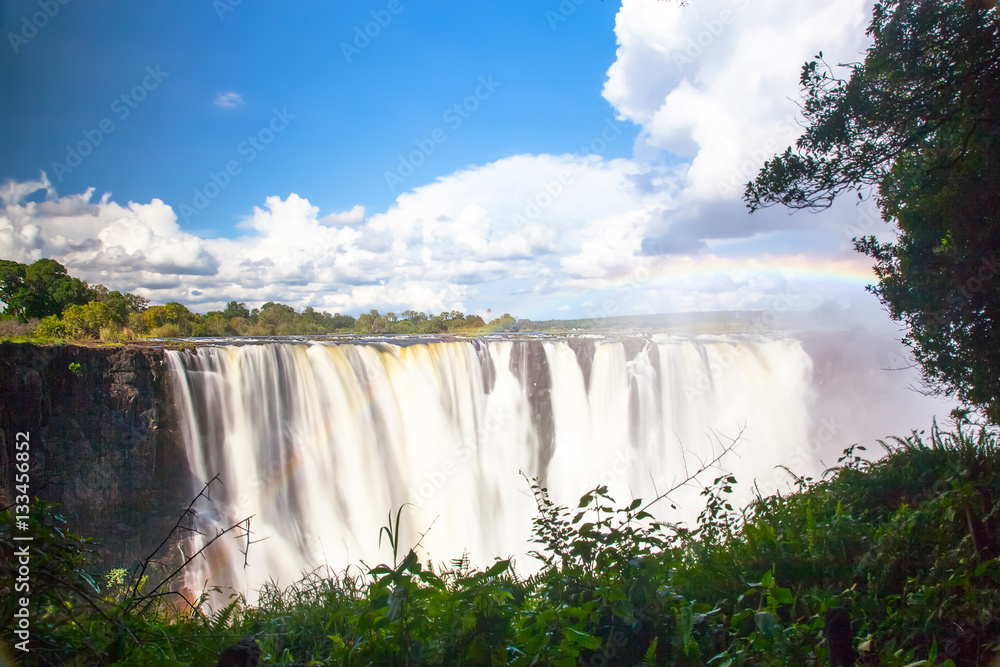 Victoria Falls.  Frontal view with a rainbow.  Taken with an MD filter.  Blue sky with clouds looming.