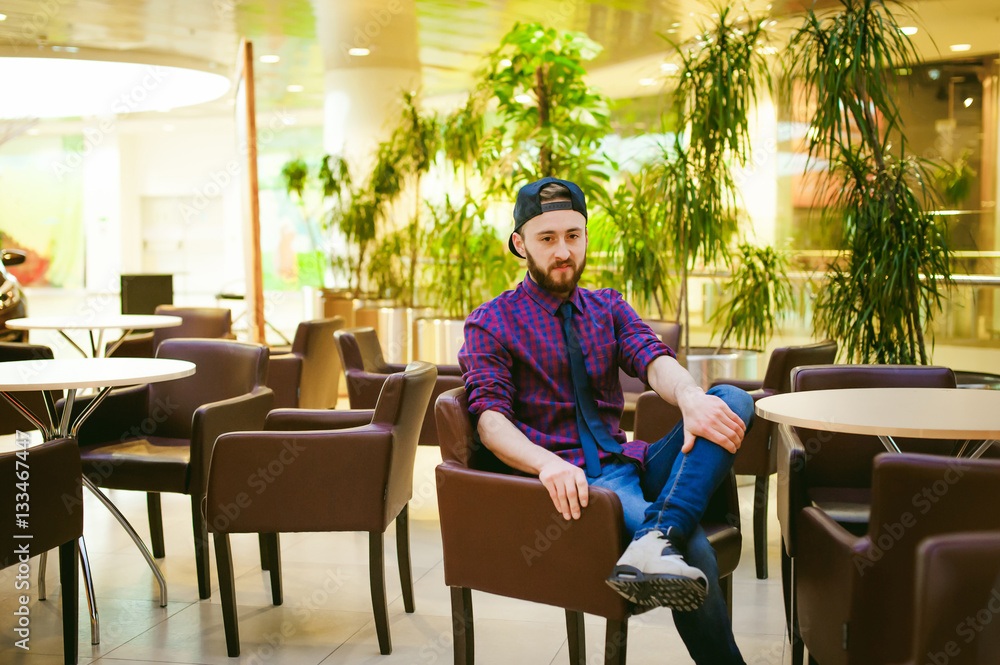 Portrait of a young man with a beard, dressed in jeans and a plaid shirt, tie and cap, sitting on a chair at the coffee shop in the mall Portrait of a young man with a beard, dressed in jeans and a plaid shirt, tie and cap, sitting on a chair at the coffee shop in the mall
