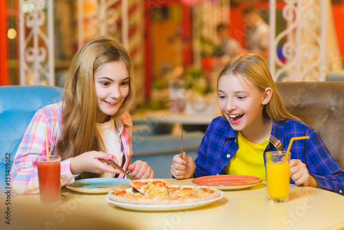Two smiling girls eating pizza and drinking juice indoor