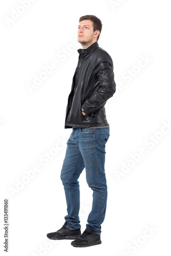 Side view of young casual man with hands in leather jacket pockets looking up serious. Full body length portrait isolated over white background.