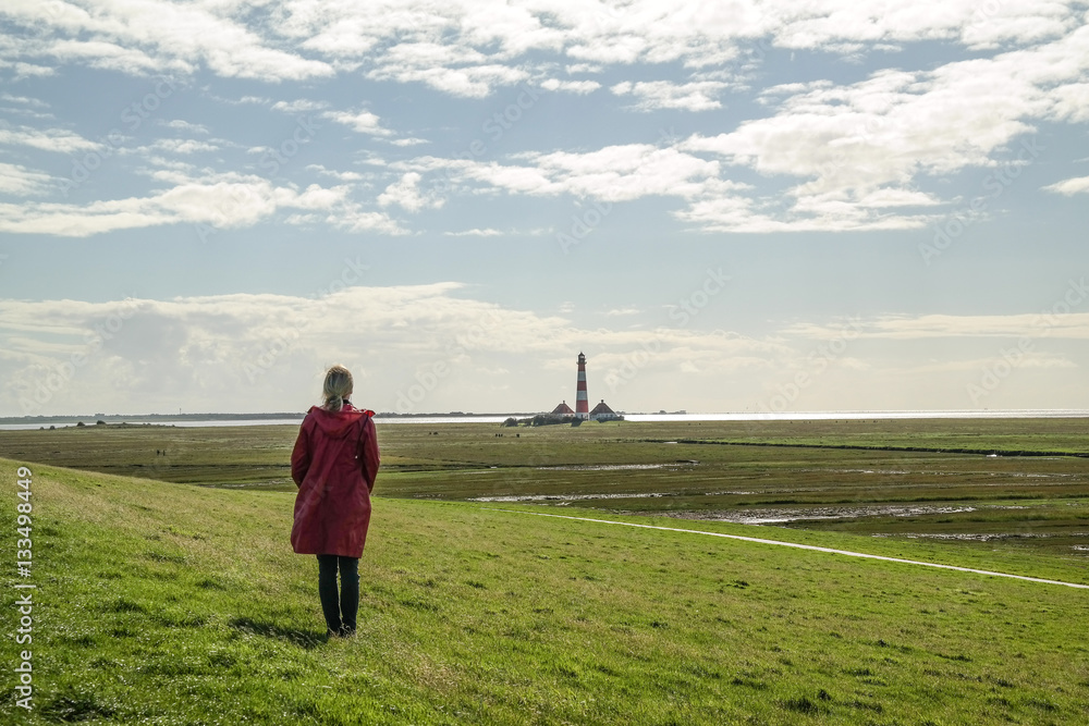 woman with red raincoat standing on green grass at north sea coast with lighthouse in distant background