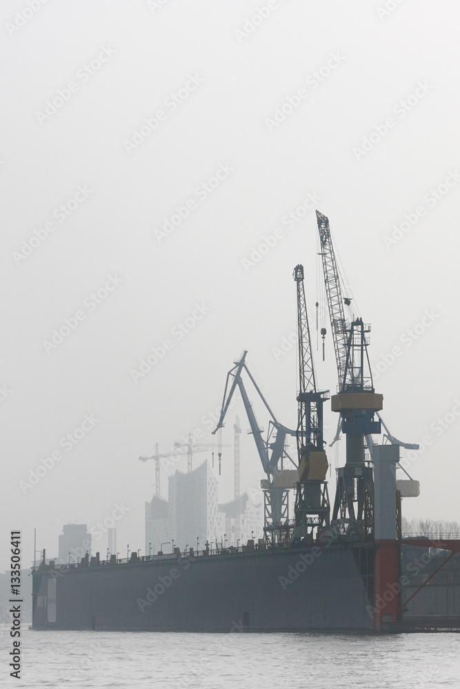 Obraz premium Floating dock in the harbor of Hamburg. Cranes of shipyard in misty morning on the Elbe river.