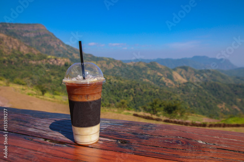 A cup of iced coffee on wood table with Mountain background.