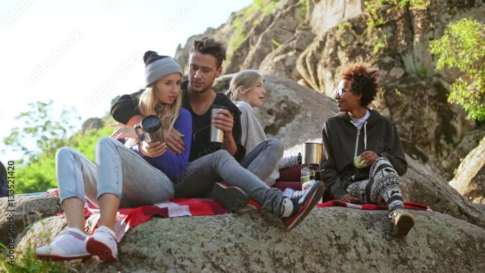 Beautiful pair sitting on plaid on rocks with thermocups while their interracial friends chatting smiling next to themin slowmotion Beautiful pair sitting on plaid on rocks with thermocups while their interracial friends chatting smiling next to themin slowmotion