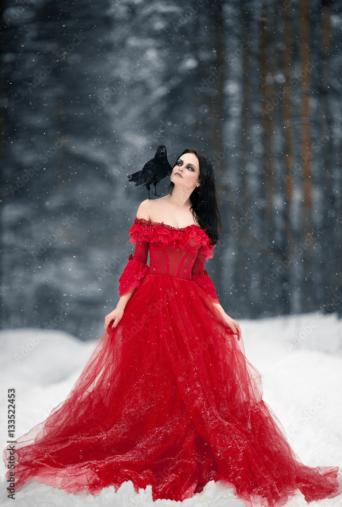 Woman witch in red dress and with raven on her shoulder in snowy