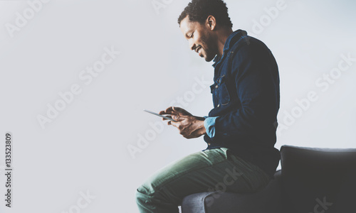 Attractive bearded African man using tablet while sitting on the sofa at his modern home office.Concept of young people working with mobile devices.Empty gray wall background.