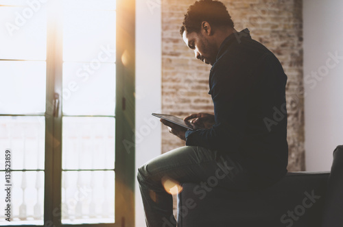 Young bearded African man using laptop while sitting on sofa at his modern home office.Concept of people working with mobile gadgets.Flares effect.Blurred window and brick wall the background.