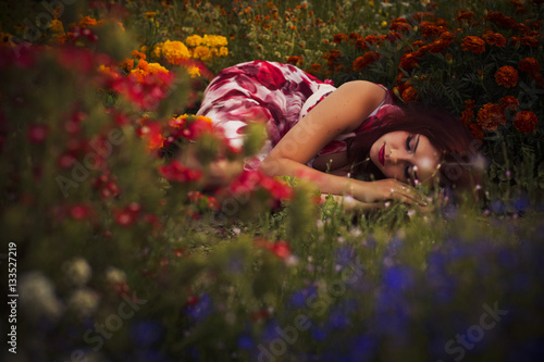 Beautiful brunette caucasian woman in white dress at the park in red and yellow flowers on a summer sunset holding flowers sitting on the grass. Copy Space