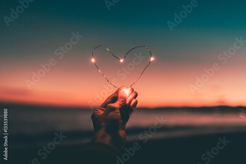 Simbolo del cuore fatto da una striscia di luci led tenuto da una mano di fronte al mare e al cielo in un tramonto rosso infuocato. San Valentino