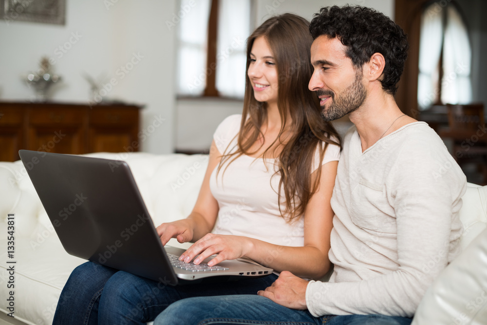 Portrait of an happy couple using a laptop computer in their house. Shallow depth of field, focus on the man