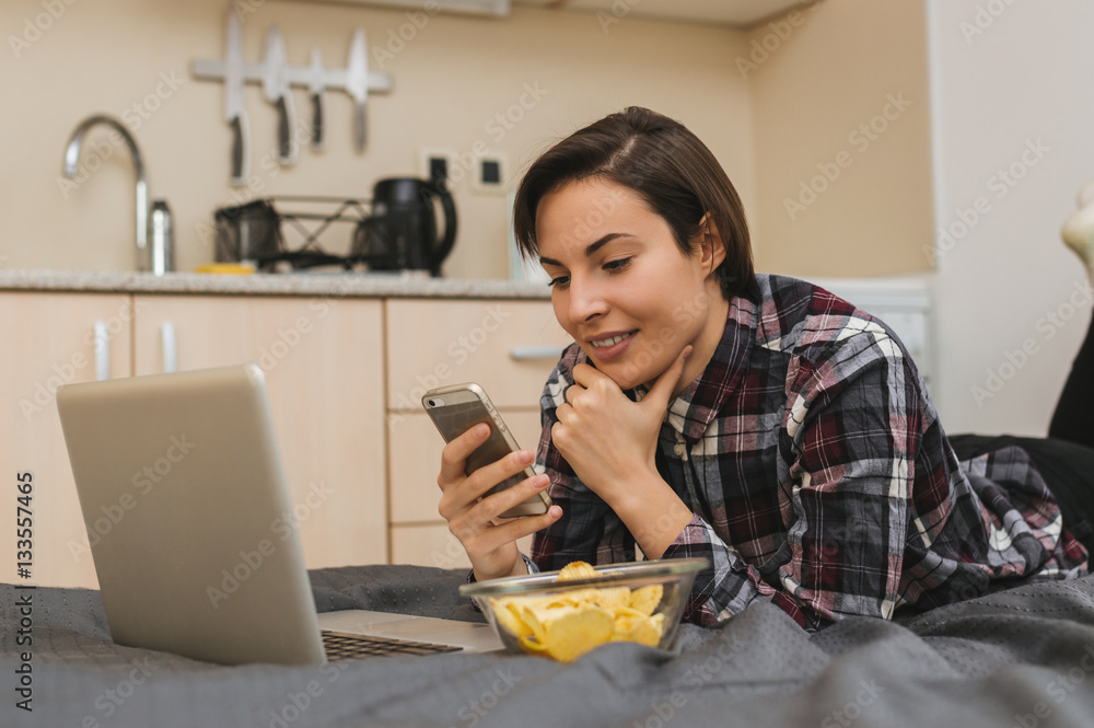 Girl on bed checking smartphone while reading news on computer Girl on bed checking smartphone while reading news on computer