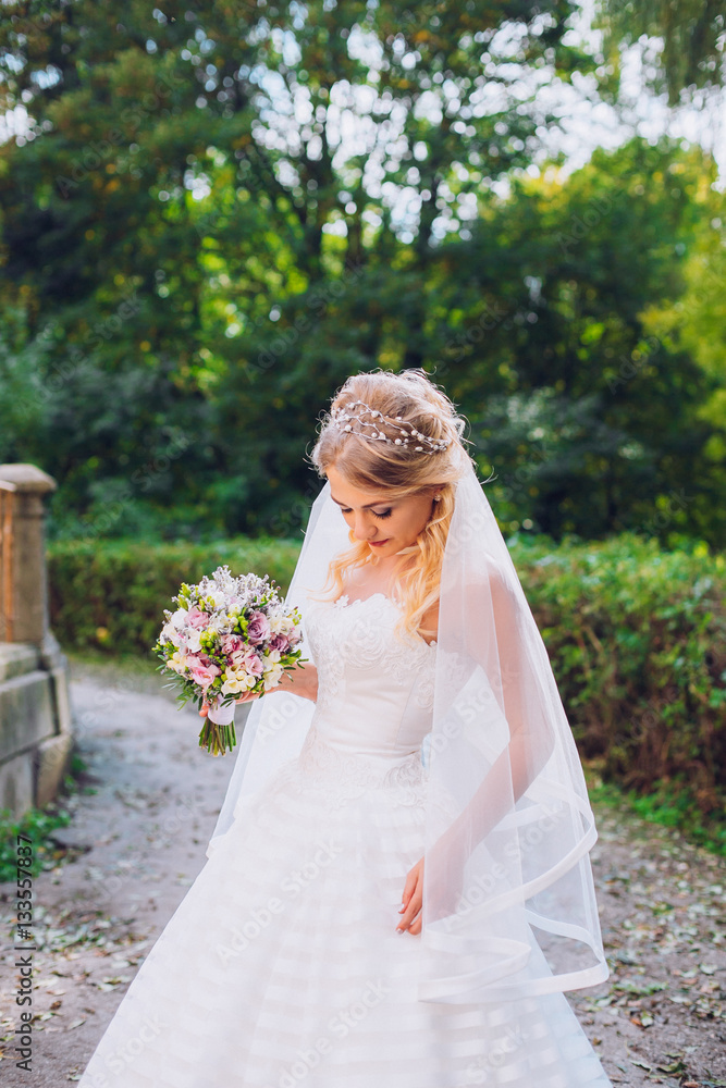Beauty bride in bridal gown with bouquet and lace veil on the nature. Beautiful model girl in a white wedding dress. Female portrait in the park. Woman with hairstyle. Cute lady outdoors Beauty bride in bridal gown with bouquet and lace veil on the nature. Beautiful model girl in a white wedding dress. Female portrait in the park. Woman with hairstyle. Cute lady outdoors