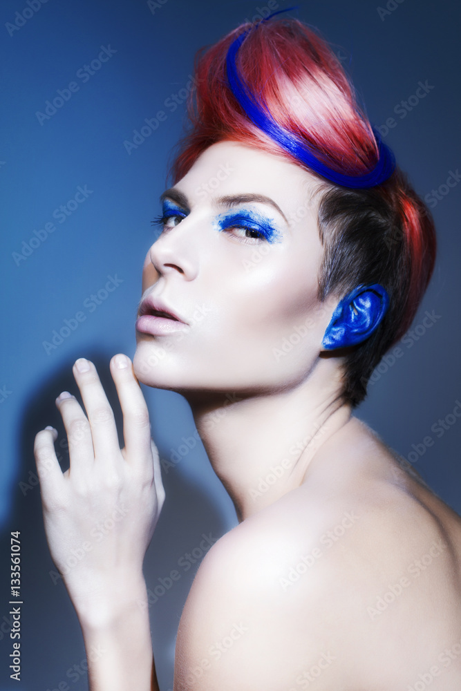 Young person with blue eye shadows, blue ears and pink hair with blue strand on it looking at camera and hands near face. Black background Young person with blue eye shadows, blue ears and pink hair with blue strand on it looking at camera and hands near face. Black background