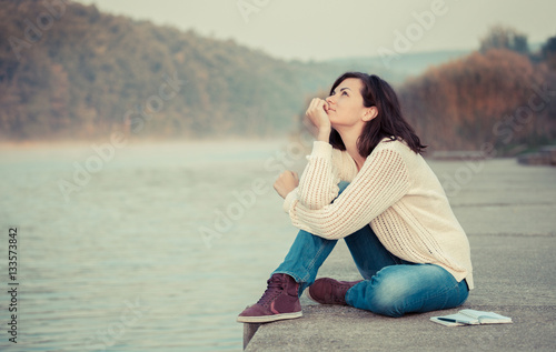 Day dreaming by lake. Young woman sitting by water and looking up in the sky.