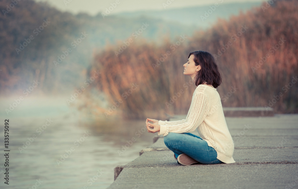 Morning meditation by lake. Young woman sitting by lake in lotus pose.