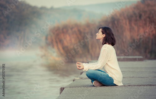 Morning meditation by lake. Young woman sitting by lake in lotus pose.