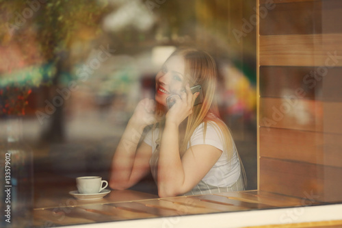 Beautiful young woman talking by mobile phone while sitting in cafe, view from outside