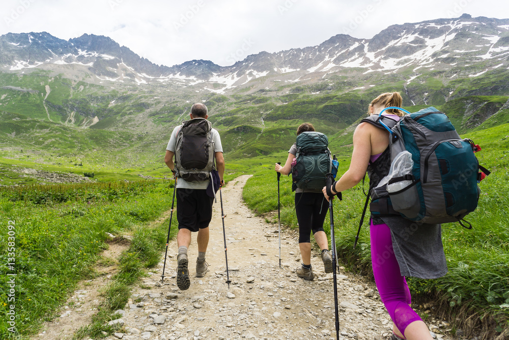 Three young hikers follow a dirt trail towards a large range of mountains in the French Alps