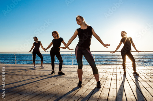 Fototapeta Naklejka Na Ścianę i Meble -  Silhouettes of sportive girls dancing zumba near sea at sunrise.
