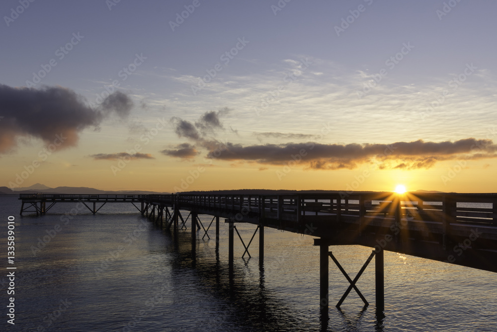 Dawn breaking at an ocean pier with a sun star,clouds and an orange glow in a blue sky
