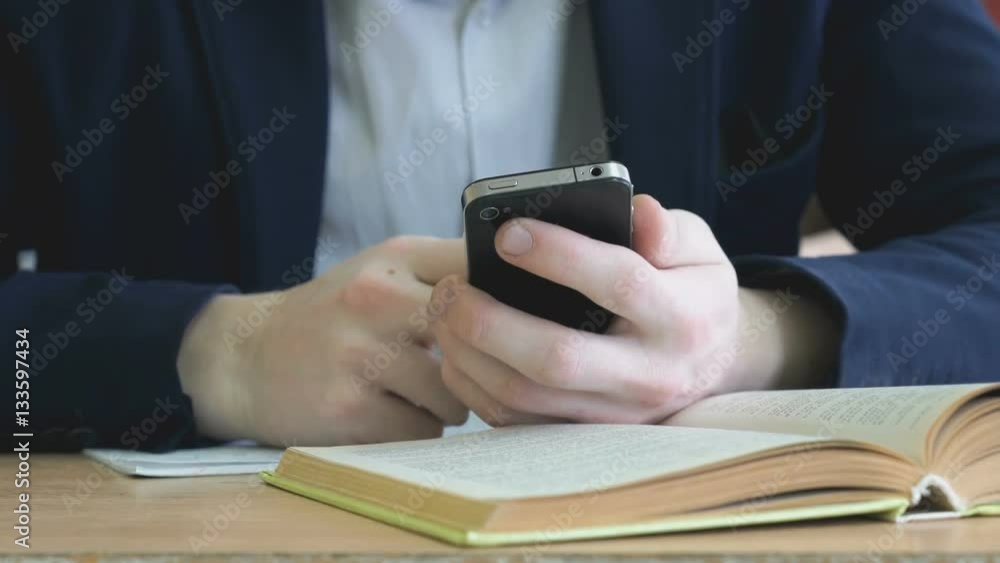 The student sitting at the school desk at school writes the text using the smartphone indoors The student sitting at the school desk at school writes the text using the smartphone indoors