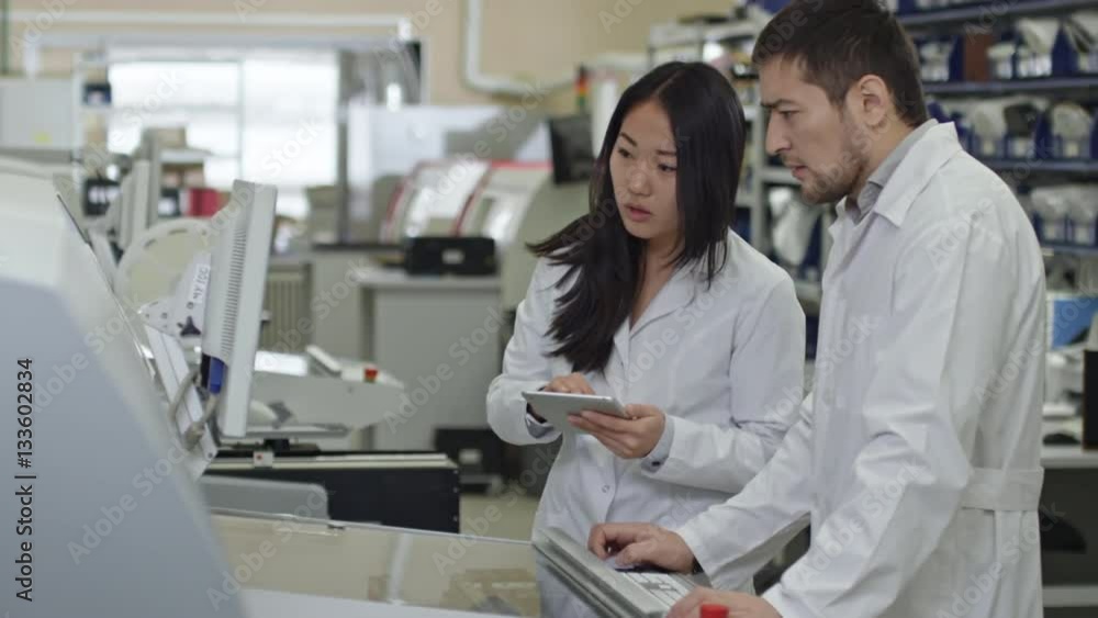 Tracking of young engineer in lab coat talking to female colleague standing at SMT machine and using digital tablet Tracking of young engineer in lab coat talking to female colleague standing at SMT machine and using digital tablet