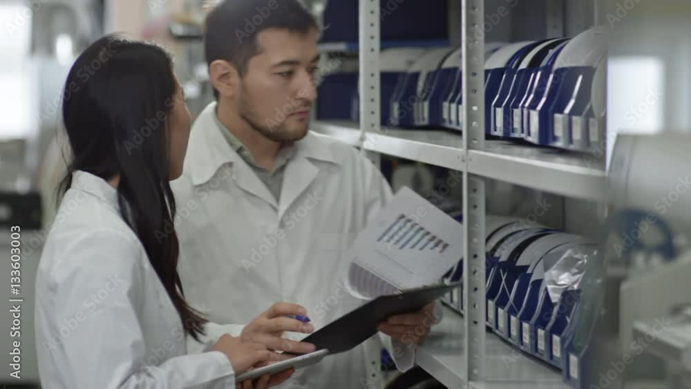 Young Asian man and woman in lab coats checking details on shelves and writing down something on documents Young Asian man and woman in lab coats checking details on shelves and writing down something on documents