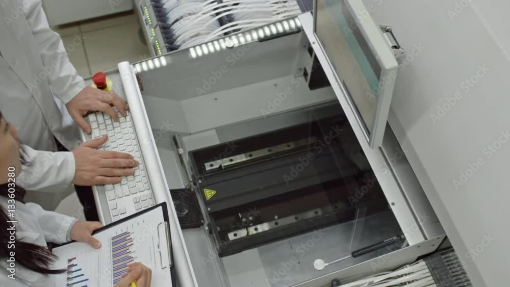 Directly above view of male engineer typing on keyboard of SMT machine while his female assistant making notes on bar graph