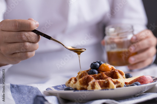 Blueberry apple waffle with honey set on a dish with a chef's hand holding spoon for a jar of honey ready to serve.