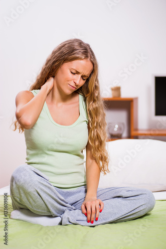 Young woman is sitting in her bed with pain in her neck.