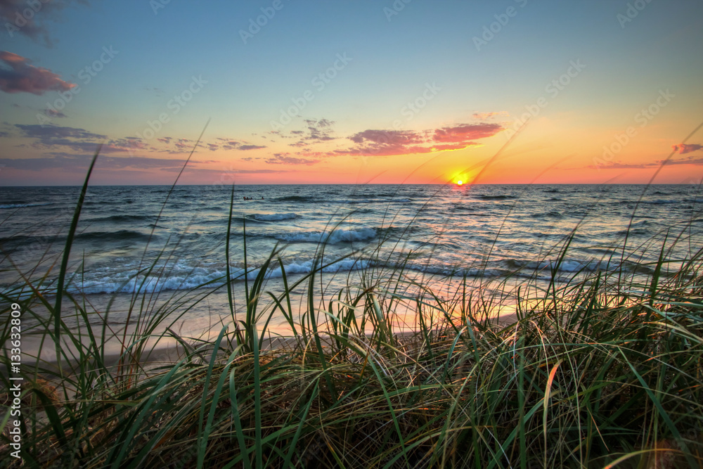 Fototapeta premium Scenic Summer Sunset Background. Beautiful sunset horizon over water with a sandy beach and dune grass in the foreground. Hoffmaster State Park. Muskegon, Michigan.