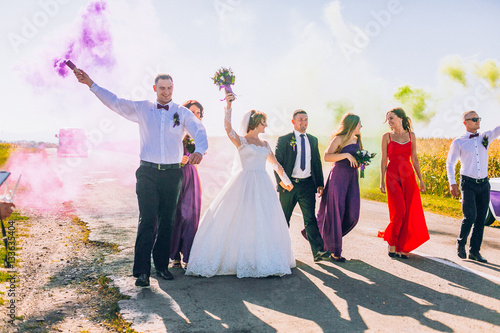 Happy newlyweds with their friends outdoor. Bride and groom with bridesmaids and groomsman walking with smoke bombs in road near retro car and yellow field.