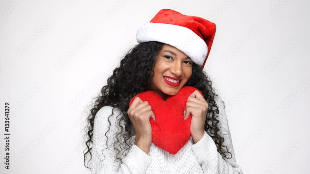 Shy African woman in white sweater and christmas hat holding plush heart in hands in studio Shy African woman in white sweater and christmas hat holding plush heart in hands in studio